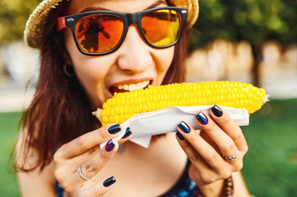 A woman in sunglasses biting into corn on the cob.